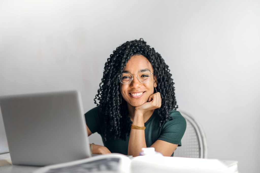 Woman sitting in front of a laptop and smiling at the camera. The woman has a textbook open beside her out of focus. She is studying or doing work at a computer.